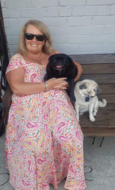 A woman in sunglasses sits on a bench, holding a black dog with a small white dog beside her.