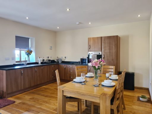 Modern kitchen with wooden dining table, chairs, and flowers; appliances and utensils visible.