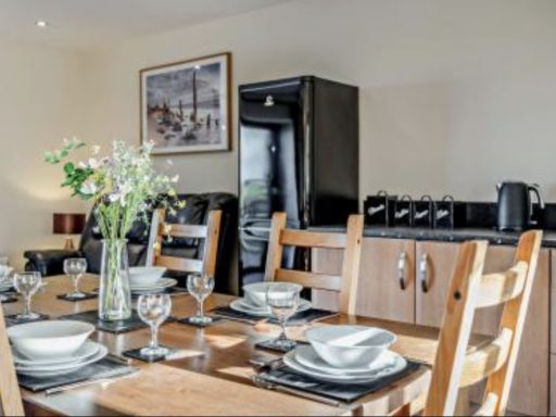 Dining area with a wooden table set for six and a modern kitchen in the background.